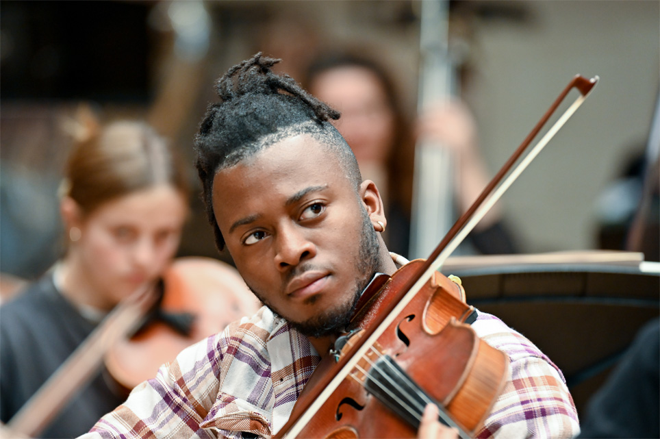 A black male student, playing the violin, rehearsing in the violin section of an orchestra.